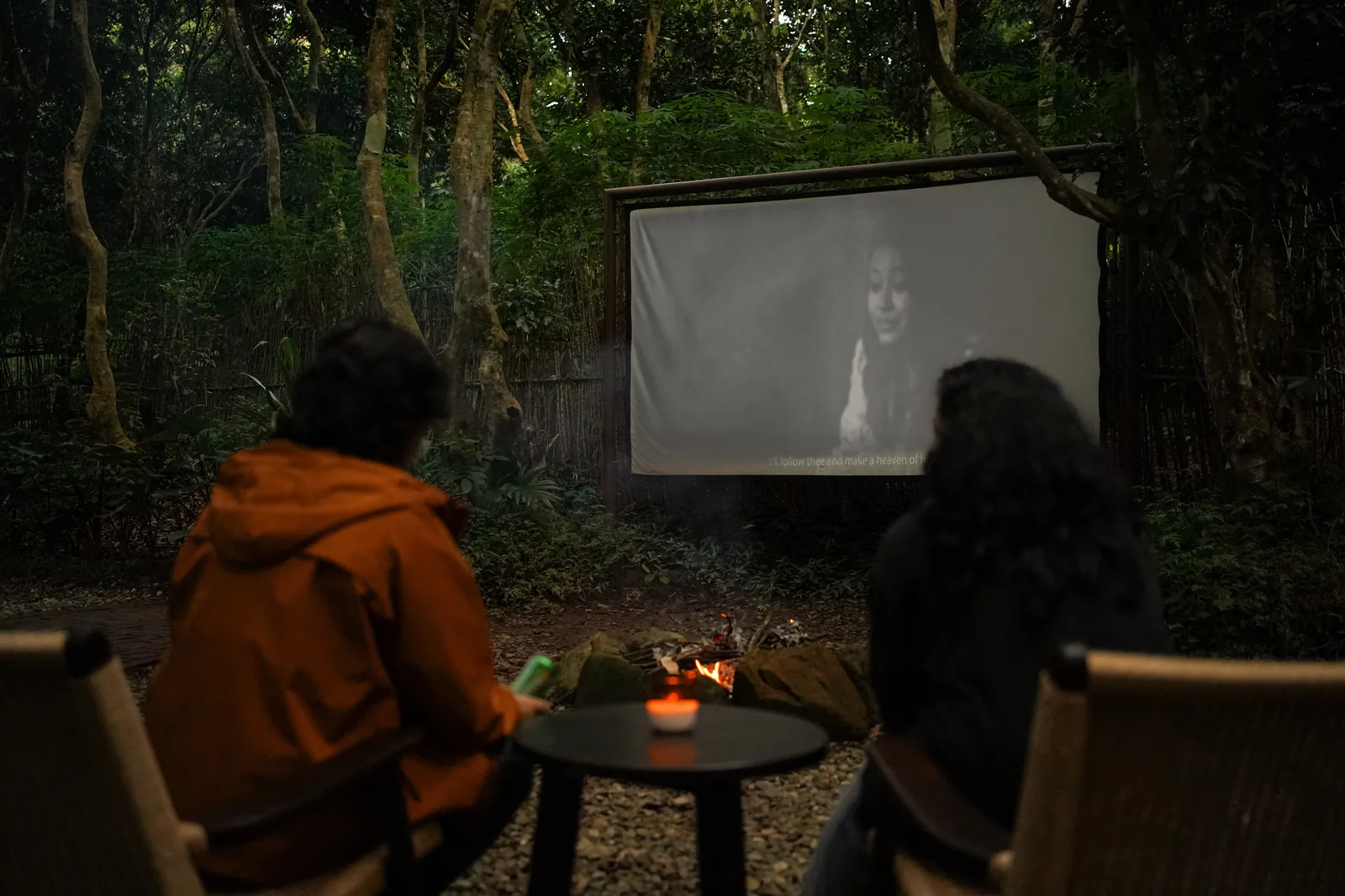 Two guests by the fire watching a film on the outdoor screen at Nan